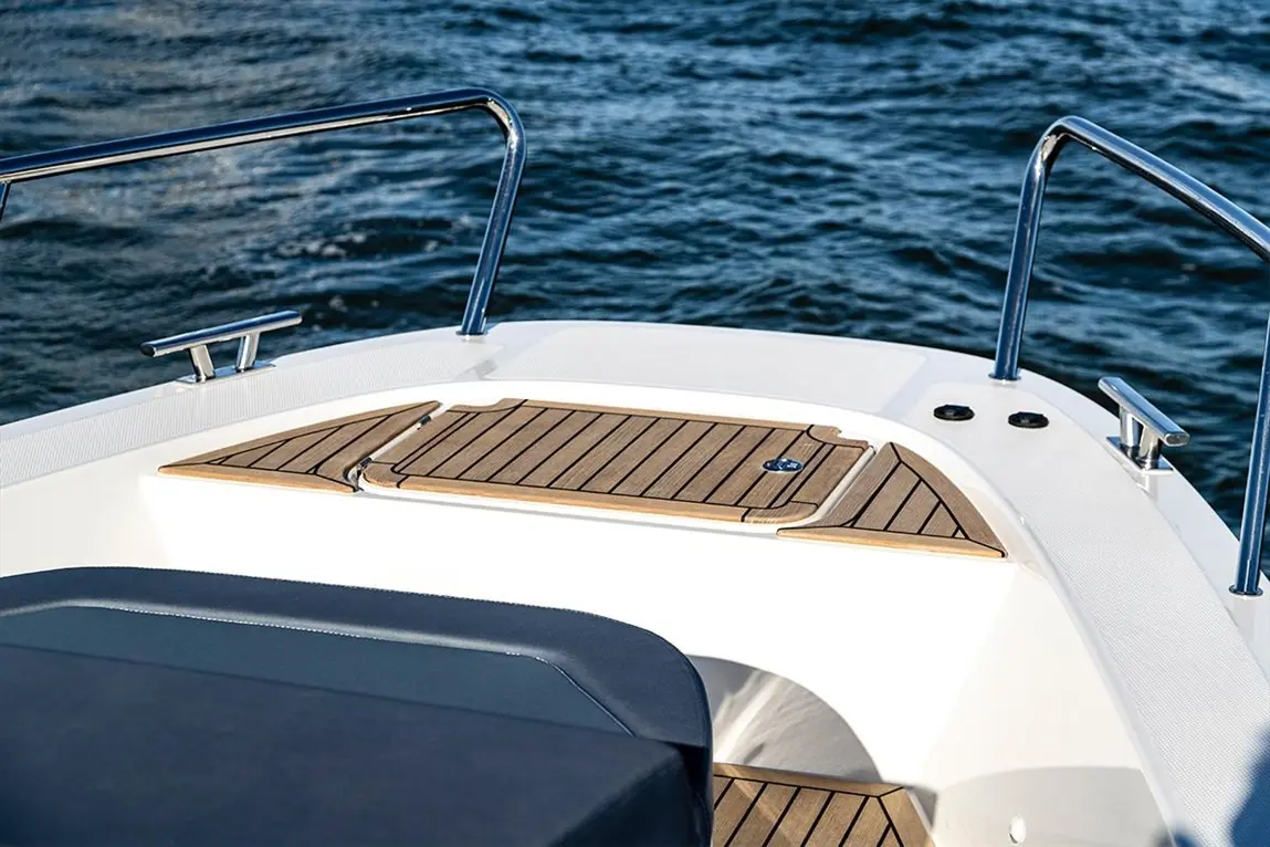 A close-up of a boat's front deck featuring wooden panels and chrome railings against a backdrop of blue water.